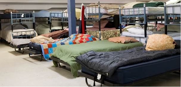 Homeless shelter with rows of basic beds and various colors of sleeping bags and pillows on top.