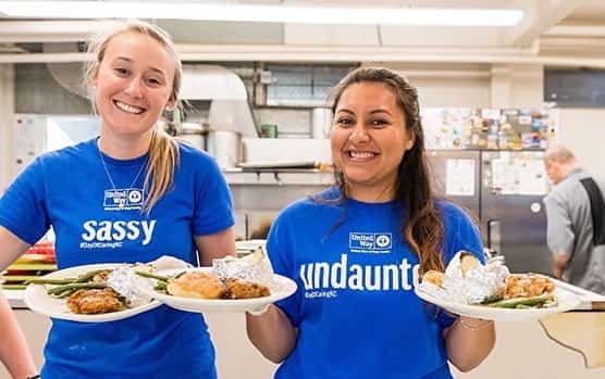 Two volunteers do holiday volunteering at a shelter or food bank, serving a meal in United Way blue Day & Night of Caring shirts.