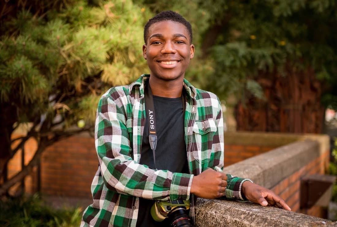 Reconnecting Youth brings out potential for opportunity youth. Image of Eddie, a black teenage male, smiling and looking forward, wearing a plaid green shirt and a high-end camera around his neck