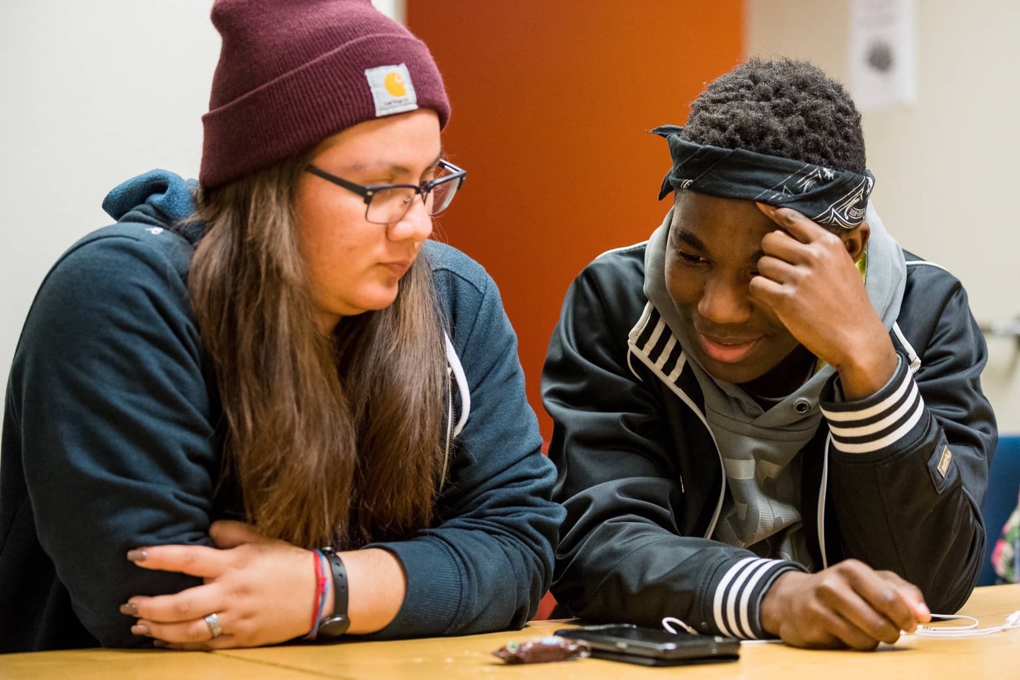 Image of a white youth female in a knit cap sitting with a black young man, both looking at a cell phone sitting on the table in front of them.