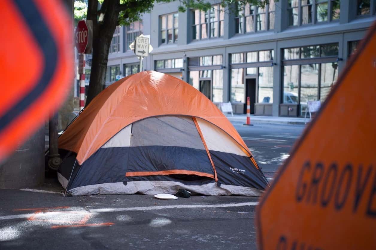 Image of an orange camping tent on a city sidewalk, flanked by street construction signs