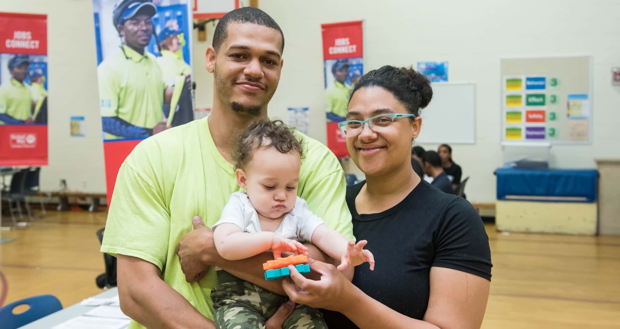 Photo of a 20-something man in a bright yellow shirt, holding a one-year-baby and a 20-something woman in green eyeglasses and black shirt with her arm around the man