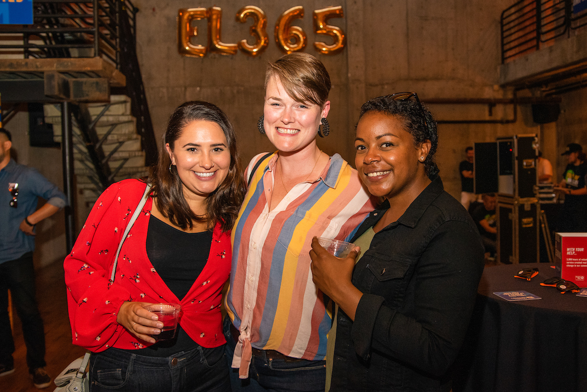 Three young professional women looking at the camera and smiling. Behind them are gold balloons that spell 