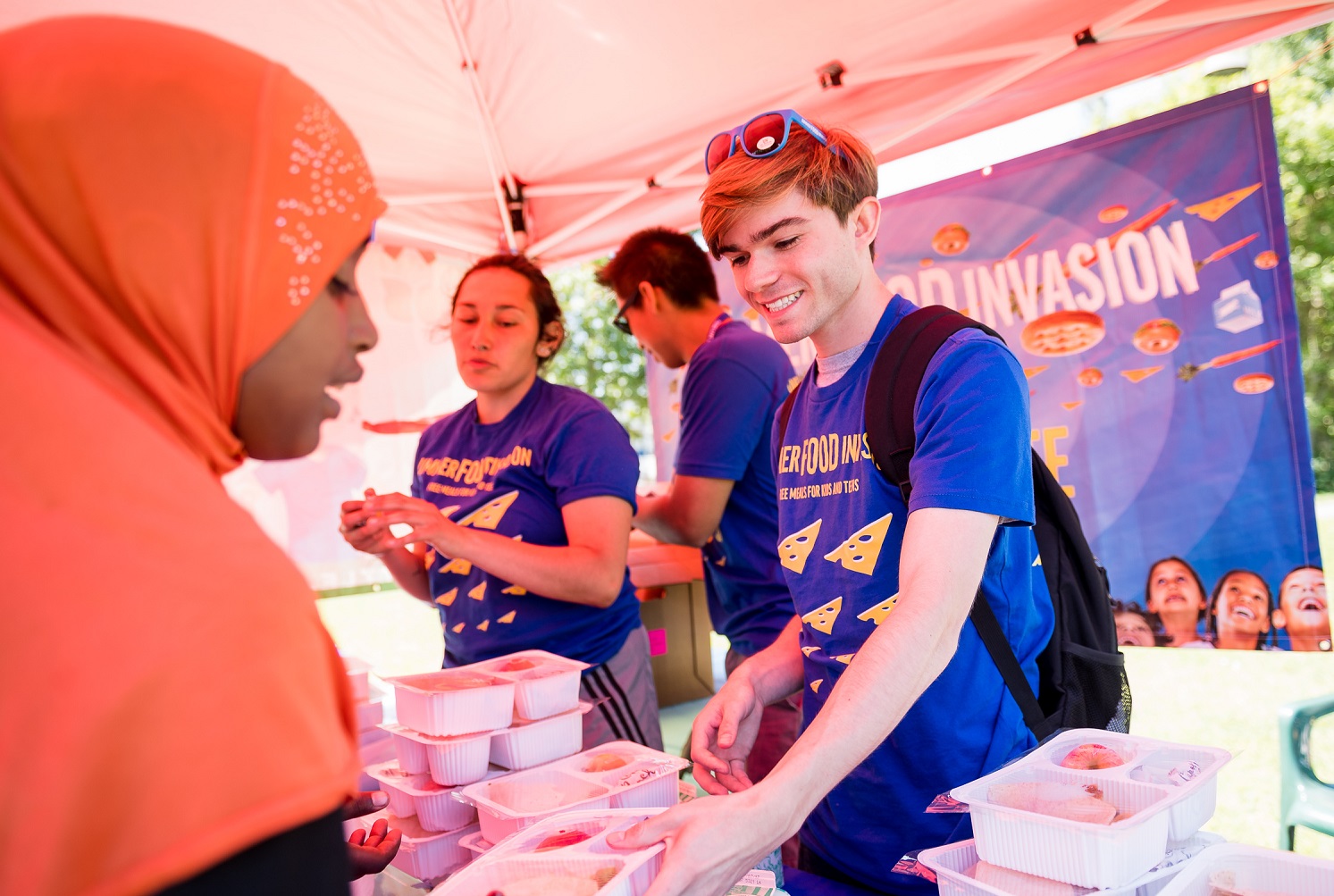 A young teen girl with an orange al-amira scarf in front of a group of smiling volunteers. One is a blond young man, handing her a plastic tray of food