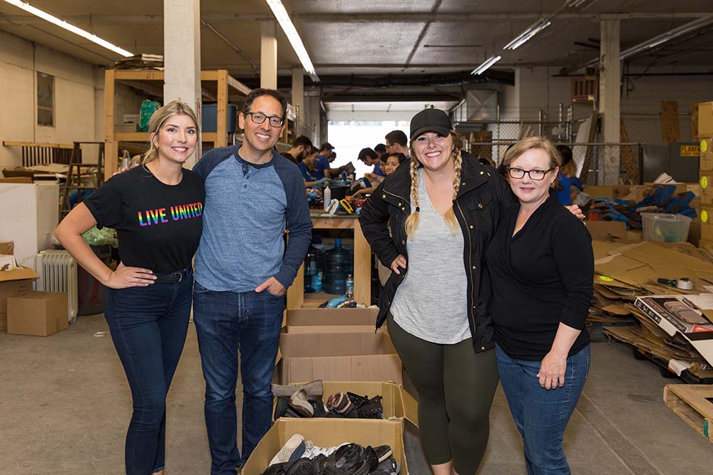 Chris and Leigh pose for a photo with day of caring volunteers in front of boxes of clothing