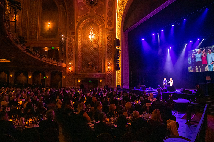 A large theatre full of people eating dinner while watching a performer speak on stage