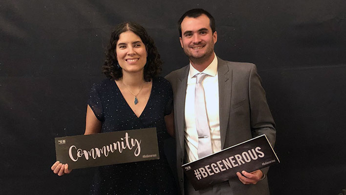 Juliana and Philip pose for a photo while holding "community" and "#begenerous" signs at the Eat, Drink and Be Generous event