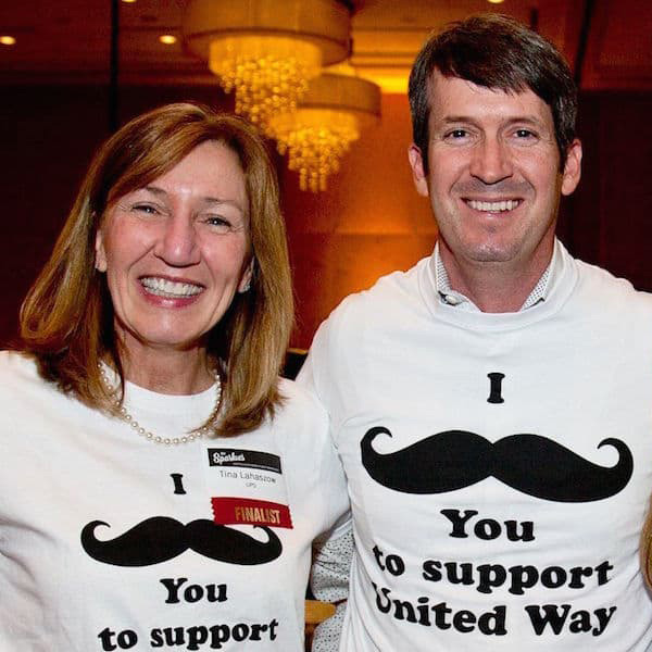 Two people stand posing for photo, both people are wearing I mustache you to support united way