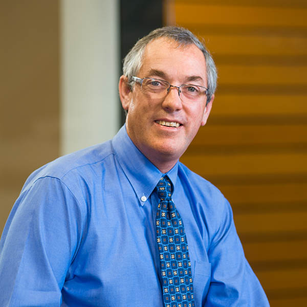 A professional headshot photo of Brian. He is sitting looking at the camera wearing a blue shirt and tie.