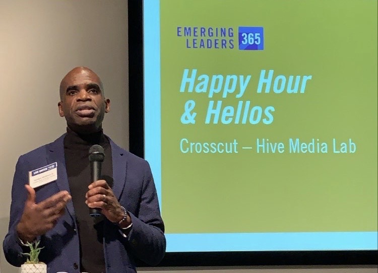 Gordon McHenry, Jr. standing in front of a large screen, holding a microphone and speaking to the attendees of the Happy Hour and Hellos Crosscut event