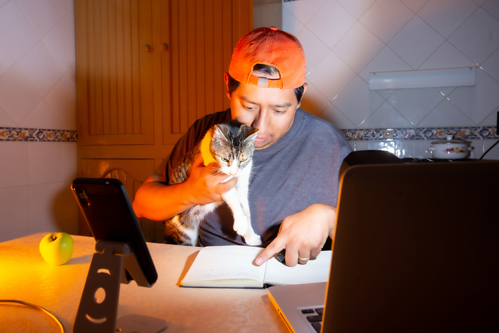 Man working at a home computer wearing a hat and holding a cat.