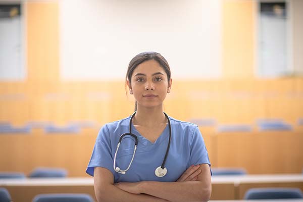 A medical student poses in a lecture hall for a portrait. She is wearing blue scrubs, has a stethoscope around her neck and her arms crossed. The student has a neutral expression on her face and has her hair pulled back into a ponytail.