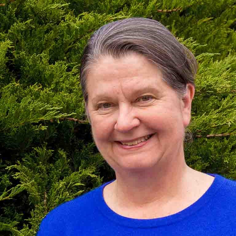 Sue Sherbrooke wearing a blue shirt standing in front of a bush and smiling