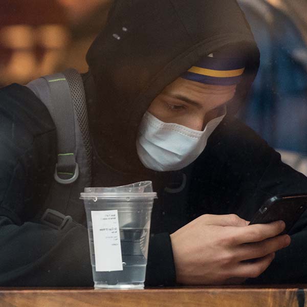 A student sits at a table in a cafe. A plastic cup sits next to the teen. The teen wears a black hoodie and is looking down at a cell phone.
