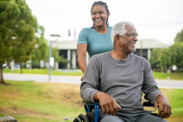 Maya pushes an older man in a wheelchair outside, they are both smiling.