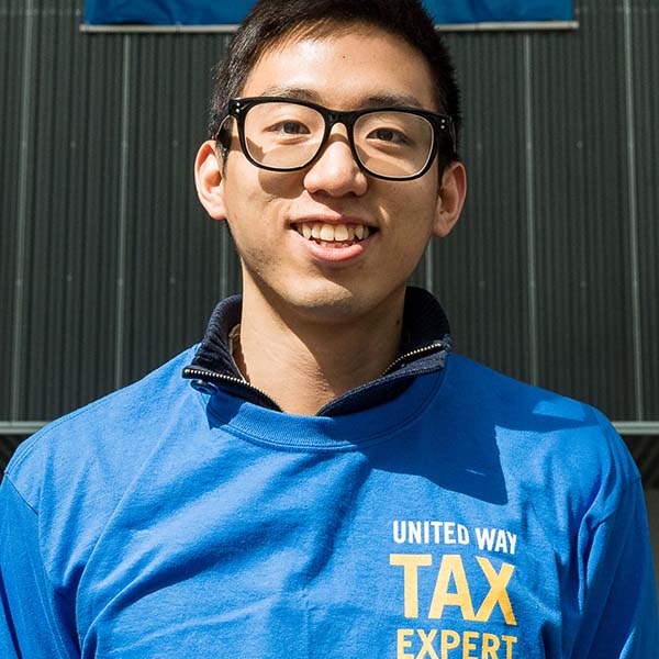 Person with short black hair, wearing glasses and a blue t-shirt that says "United Way Tax Expert" smiles toward camera