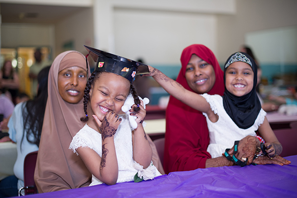 Two adults wear traditional Muslim head coverings sit next to each other. They both have a young child sitting in their laps. One child is also wearing a head covering and smiles while she reaches out to touch the graduation cap worn by the other child. 