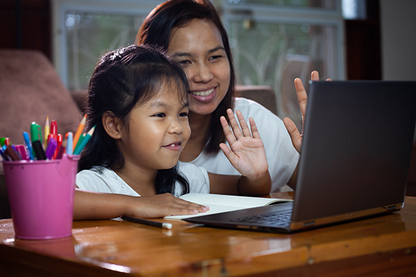 A young child and and adult both sit and have their left hands raised while smiling and looking into a laptop monitor during an online learning session.