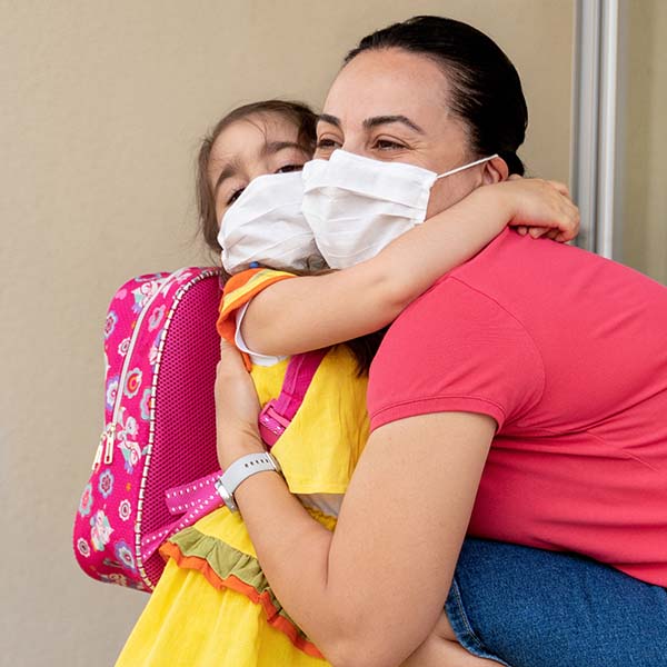 A woman kneels down to hug a young child. The young child is wearing a backpack. Both are wearing masks during as safety precaution for the COVID-19 pandemic.