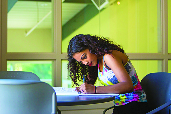 Student sits at a table in a classroom, is looking down and writing on a piece of paper.