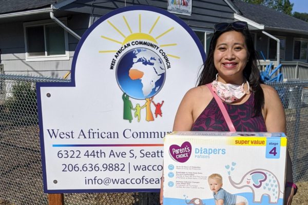 Tiffany stands in front of the West African Community center, smiling and is holding a box full of diapers.