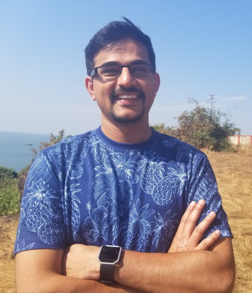 Anand Naik stands outside with arms crossed while looking at the camera and smiling. He wears glasses, and a blue shirt with a firework pattern.