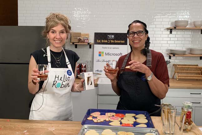 Robin Wehl and Donna Moodie stand in a kitchen behind a counter with a tray full of cookies, while smiling and holding up cocktails.