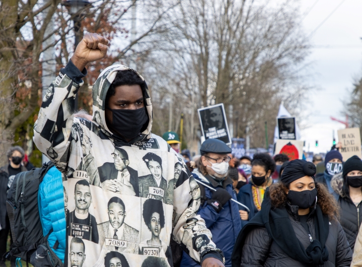 A man standing at a Martin Luther King Day march with his right fist in the air wearing a jacket with photos of Black historical figures.