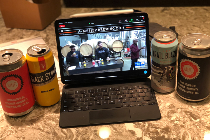 A laptop sits on the counter displaying the Eat, Drink and Be Generous event. Two beers from Metier Brewery sit on either side of the laptop