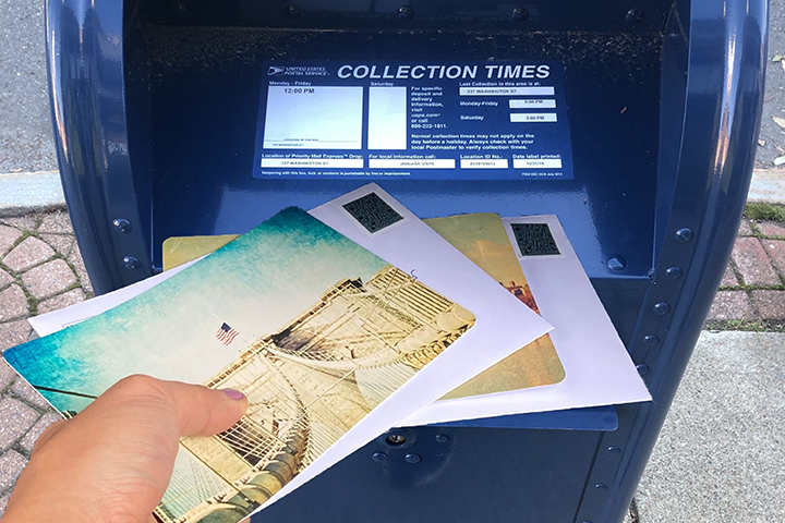 A hand inserts several envelopes into an outdoor USPS mailbox.