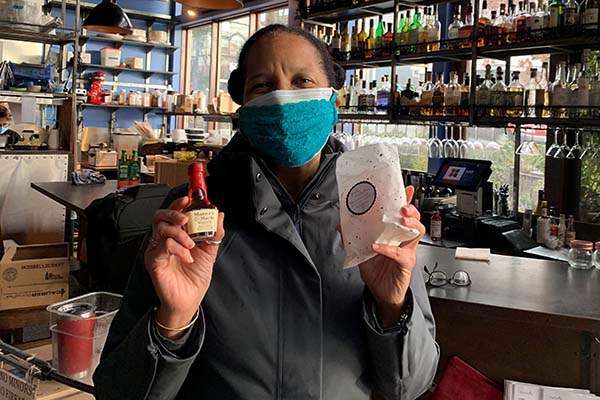 Donna Moodie, owner of Marjarie, is wearing a teal mask inside her restaurant and is holding a mini bottle of makers mark in one hand and a bag of cocktail supplies in the other.