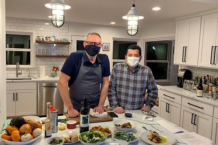 Ethan Stowell and Fred Rivera wear face masks and are standing behind a table with several ingredients prepped for a cooking demonstration