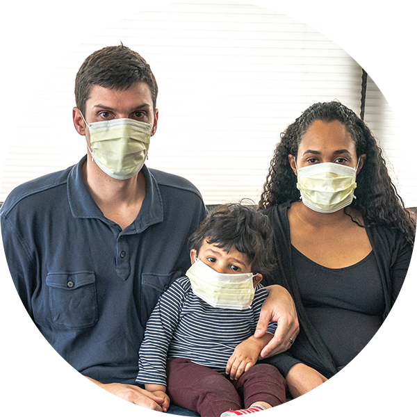 Two adults sit on a couch with their child sitting between them. The father has his arm around the child's shoulder. All three of them wear medical face masks