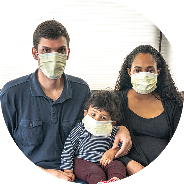 Two adults sit on a couch with their child sitting between them. The father has his arm around the child's shoulder. All three of them wear medical face masks