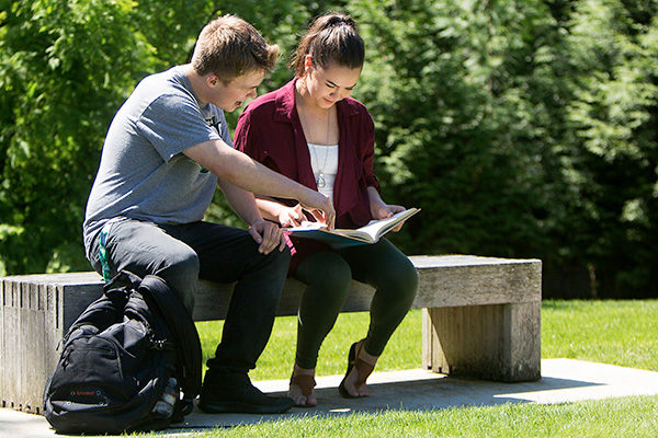 two students sit on a bench outside, they are looking down at an open book, one student points down toward a page.