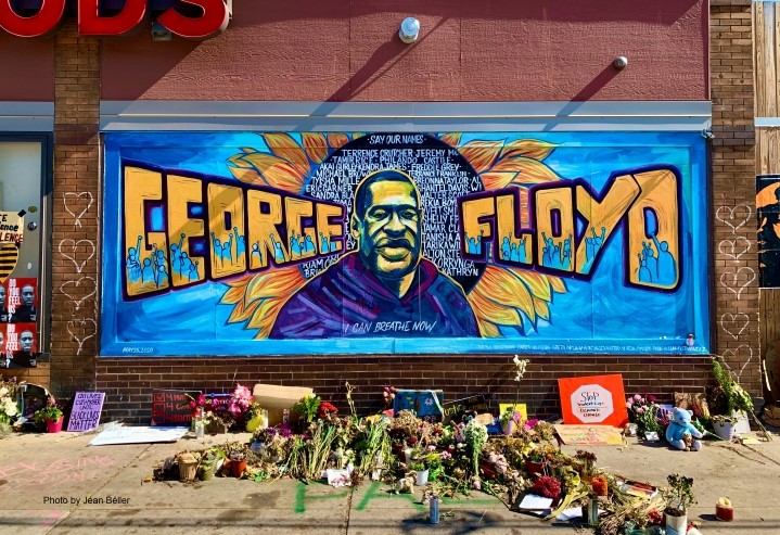 A mural in Minneapolis with a painting of George Floyd and his name painted next to him. Flowers are on the ground below the mural.