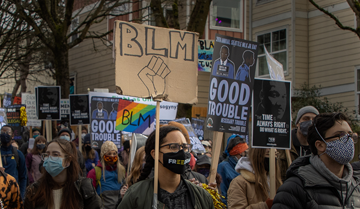 Several people wearing face coverings at a Martin Luther King Jr. Day march holding BLM signs and other posters on wood sticks.