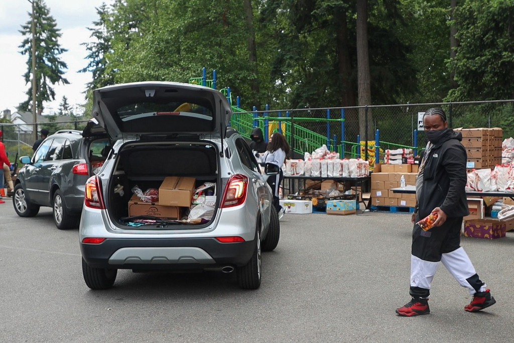 A man wearing a facial covering helping to load a package of food into the open trunk of a car that shows more food already loaded. There are tables with packaged foods on tables in the background.
