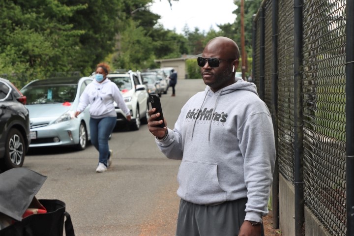 A man wearing sunglasses holding a mobile phone wearing a sweatshirt. In the background a woman wearing a mask is walking along a line of cars.