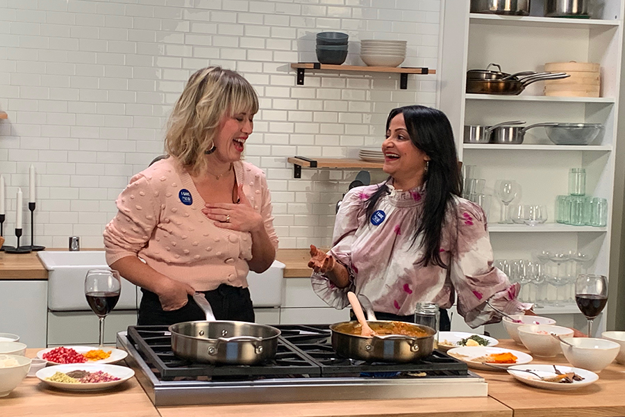 Brook and Preeti from an eat dring and be generous event stand in front of a stove with pots of food cooking looking at each other and laughing.