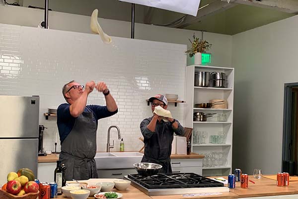 Ethan Stowell and Manny Chao are in a studio kitchen. Ethan is demonstrating how to flip a pizza into the air while Manny watches. 