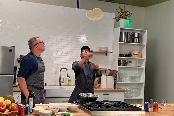 Ethan Stowell and Manny Chao are in a studio kitchen. Manny is flipping a pizza into the air while Ethan watches.