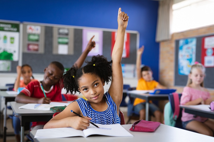 African american girl raising her hands while sitting on her desk in the class at school. school and education concept