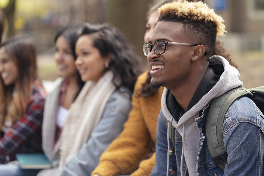 A middle-sized group of university-aged students, both male and female, are sitting outside on the campus grounds and relaxing before class. They are dressed in casual but stylish clothing while smiling.