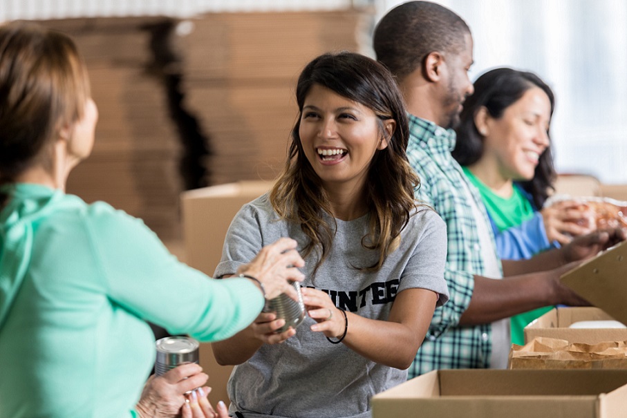 Cheerful mid adult Hispanic female volunteer accepts a canned food donation during community food drive. She is wearing a gray volunteer t-shirt. Her friends are volunteering in the background.