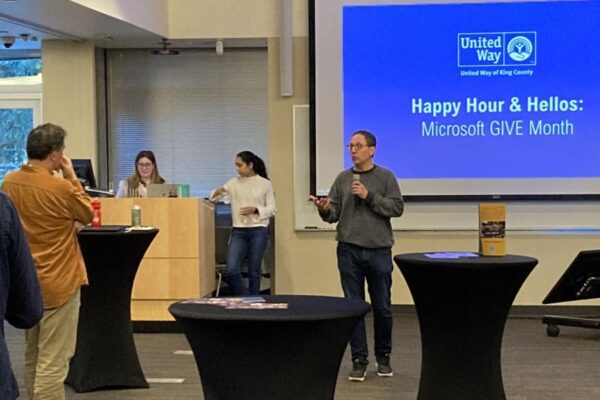 Gordon McHenry, Jr. standing in front of a large screen, holding a microphone and speaking to the attendees of the Happy Hour and Hellos Crosscut event