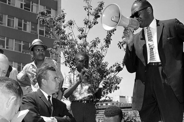 A man standing at a Martin Luther King Day march with his right fist in the air wearing a jacket with photos of Black historical figures.
